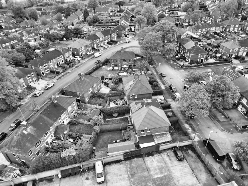 A high-angle black-and-white aerial photograph of a suburban residential neighbourhood showing multiple houses with varied roof styles, front and back gardens, and parked cars along the streets. In the foreground, several houses with fenced gardens and small outbuildings are visible, with some gardens containing trees and shrubs. The streets are lined with vehicles parked parallel, and a few moving or stationary cars can be seen. The background features a larger road with more parked cars and houses, as well as a few larger trees providing greenery amidst the housing. The image depicts a typical UK residential area, suitable for home relocation and furniture transport services, and highlights the spatial arrangement relevant to loading and unloading processes for professional removals by companies such as Man with Van Lampton.
