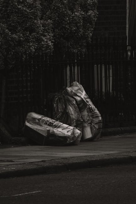 Two large, crumpled black plastic bags filled with household waste are piled on a paved pavement next to a black metal fence with vertical bars and a brick wall behind it. The bags contain paper, cardboard, and plastic packaging materials, visible through tears in the plastic. The scene is outdoors with natural lighting, suggesting a residential area. This image illustrates waste disposal or clearance activities related to home relocation or moving services, such as those provided by Man with Van Lampton, supporting the context of adhering to local bulky item rules for residents. No equipment or vehicles are visible in the scene.
