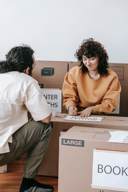 Indoor scene showing several cardboard boxes of varying sizes arranged on a table near a window with yellow curtains. One box is sealed with clear packing tape, while others are open or partially cove