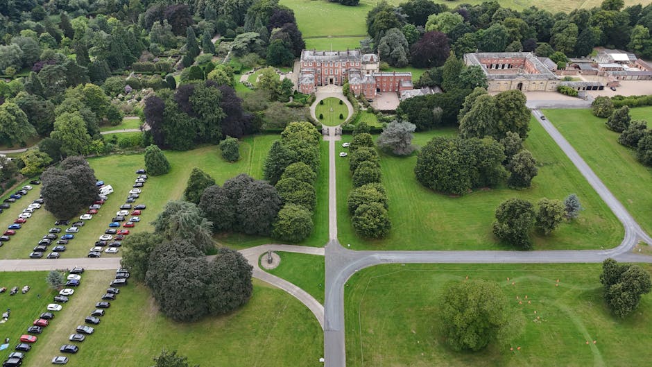 An aerial view of a large estate with a historic red-brick building at the top center, surrounded by well-maintained green lawns, pathways, and numerous mature trees. To the left, a spacious parking area filled with rows of parked vehicles is visible, with some small pathways leading through the landscaped grounds. In the foreground, there is a driveway intersection and a curved pathway that connects to a gravel or paved driveway running along the right side of the estate. The scene shows no moving activity but highlights the accessible outdoor environment ideal for house removals or furniture transport, with clear, natural lighting demonstrating the layout of the property and its surrounding grounds, as managed by services like Man with Van Lampton as part of a home relocation or packing and moving process.