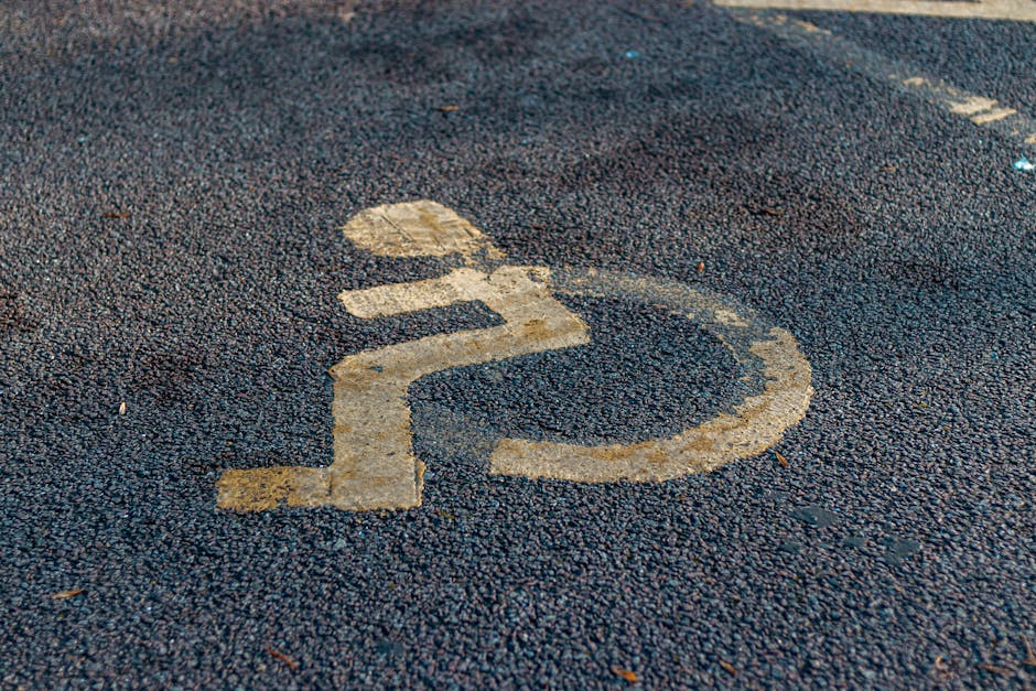 Close-up view of a worn yellow wheelchair accessibility symbol painted on a textured dark asphalt surface, indicating designated parking for individuals with disabilities outside a residential property. In the background, there is a driveway with a white van and a stack of cardboard boxes, some wrapped in plastic, being loaded or unloaded near the entrance of a house on Lampton Park Estate. The scene suggests a home relocation or furniture transport process facilitated by a removals service, with visible equipment such as trolleys and blankets used for safe handling of household items. Natural daylight illuminates the area, highlighting the details of the moving activity and the surrounding environment, emphasizing the importance of accessible parking during house removals conducted by Man with Van Lampton.