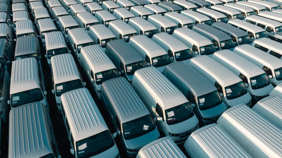 An aerial view showing a large parking area filled with numerous white vans, all parked in orderly rows on a paved surface. The vans are identical, with ribbed metal roofs and large windshields, refle