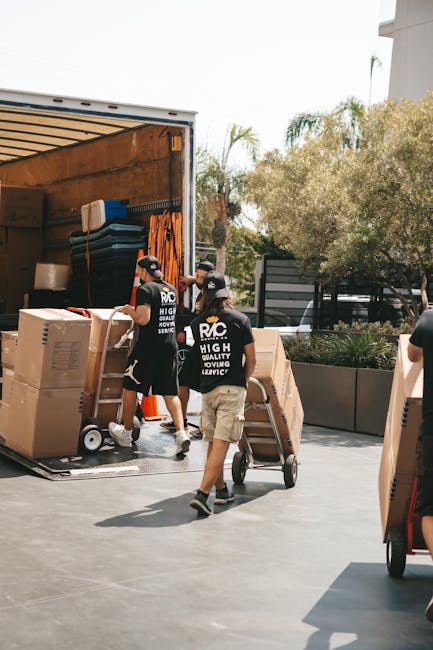 Two movers from Man with Van Lampton are engaged in a home relocation, loading large cardboard boxes and furniture onto a moving truck situated outdoors on a paved area. One mover, wearing a black T-shirt with the company's logo and beige shorts, is pushing a hand truck loaded with a piece of furniture or a box, while another mover, also in a black T-shirt with the logo, stands inside the truck arranging packed items and equipment such as black luggage, plastic containers, and orange straps. Additional boxes and packing materials are visible inside the truck, which is open to facilitate the loading process. The scene is set against a backdrop of trees, a building with modern architecture, and a partly cloudy sky. The photograph captures the organized packing and furniture transport phase typical of house removals, reflecting the logistics and handling involved in professional moving services provided by Man with Van Lampton.