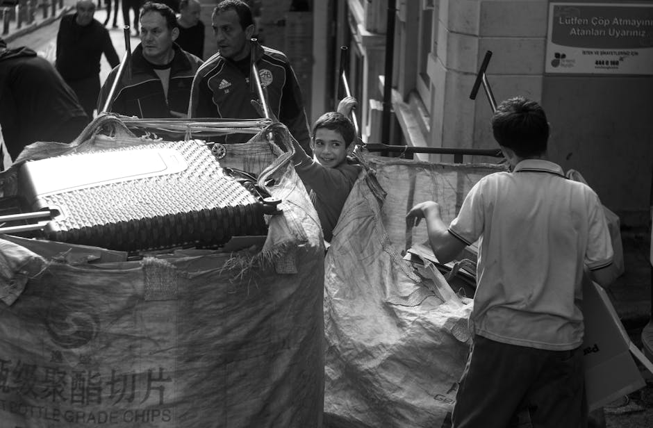 The image displays a group of people involved in a home relocation process, with a focus on furniture and packaging materials. In the foreground, a large, partially open cardboard or fabric-covered container holds various household items, including a plastic-wrapped appliance and a foam mattress or padding wrapped in plastic. A smiling child is visible inside the container, gazing toward the camera. Surrounding the container, several adults are engaged in packing or loading activities; one person wears a light-coloured collared shirt and appears to be organizing or carrying a box, while others in the background are handling larger items or observing the scene. The environment suggests an urban setting, possibly on a street or driveway adjacent to a building, with a building wall and a sign visible on the right side of the image. A vehicle, likely a van used for removals, is positioned nearby, hinting at professional furniture transport or moving services. The lighting is natural, indicating daytime, and the overall scene captures the multi-person effort involved in a household move, with clear attention to packing, lifting, and loading activities related to home relocation.