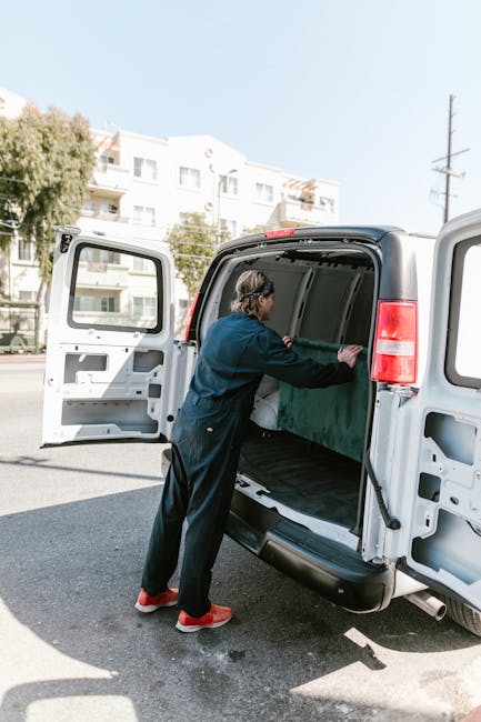 A young man with light brown, wavy hair wearing a black bandana and a navy blue jacket is sitting in the driver's seat of a white moving van. He is smiling and giving a thumbs-up gesture through the o