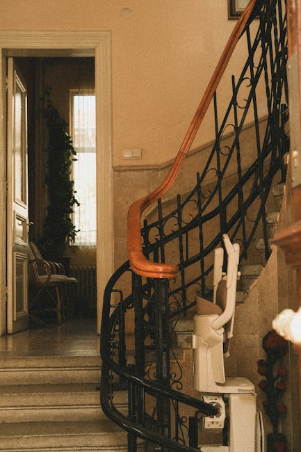 Inside a residential property during a home relocation process, a staircase with a black wrought iron balustrade and wooden handrail leads to an upper floor. At the base of the stairs, a white stair chair lift is installed, indicating accessibility adaptations for moving residents. To the left, an open doorway reveals a room with a large window allowing natural light to illuminate a beige wall and a vintage-style wooden bench with fabric upholstery positioned beside a decorative cabinet. Cardboard boxes and packing materials are not visible in this section of the image. The environment suggests an organised indoor space prepared for furniture transport and packing during the moving process, with lighting highlighting the staircase, lift, and surrounding interior features, as managed by professionals such as Man with Van Lampton.