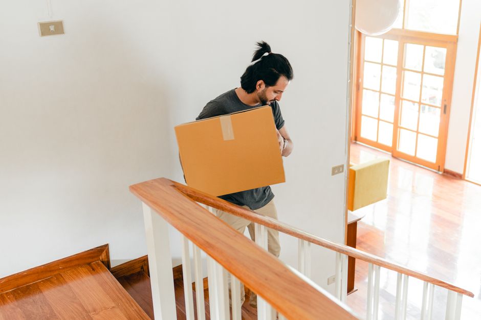 A man with dark hair and a beard, dressed in a grey t-shirt, is carrying a medium-sized cardboard box wrapped with clear tape over his shoulder while walking down a staircase inside a house. The staircase has a wooden handrail and white balusters, and is situated in a well-lit interior with natural light coming through large glass doors with wooden frames at the bottom. In the background, there is a small piece of furniture and some packaging materials. The scene depicts the household moving process, with the man from Man with Van Lampton involved in preparing belongings for home relocation or furniture transport on the second floor of a Lampton flat, as part of a professional removals service.