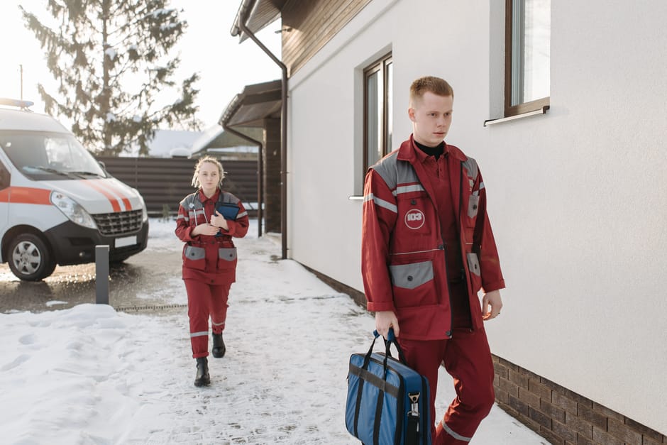 Two emergency medical responders wearing red uniforms with reflective grey stripes are standing outside a residential building during daylight. The male responder in the foreground is holding a blue medical bag in his right hand and looking to the side, while the female responder in the background is carrying a tablet or clipboard under her left arm. Both are on a snow-covered pathway beside the house, with the house's white exterior wall and a window visible behind them. To the left, a white and orange emergency vehicle is parked on a paved area near the house, with trees and a fence in the background. The scene depicts a calm moment during a home relocation or emergency response, with the responders prepared with medical equipment in an environment with natural daylight. This image is relevant to the services offered by Man with Van Lampton, especially in the context of house removals and moving logistics in urgent situations.