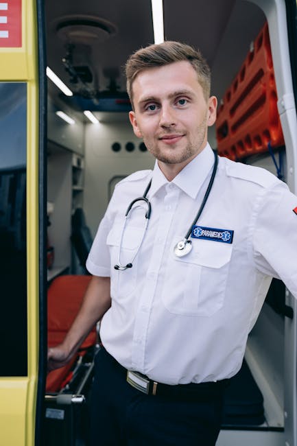 A young male paramedic with light skin, short brown hair, and a friendly expression, wearing a white uniform shirt with a stethoscope draped around his neck, standing inside the open doorway of an ambulance. The ambulance's interior, which includes medical equipment and seating, is visible in the background. The ambulance has yellow exterior panels, with the man positioned near the entrance, ready to assist or transport patients. This scene is related to emergency medical services, which can be a part of urgent moving or relocation support as offered by Man with Van Lampton during home relocation or urgent moves involving medical needs.
