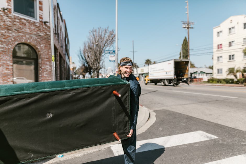 A man with a bandana on his head is seen walking along a street curb while carrying a large, rectangular mattress or padded furniture piece covered in dark fabric with green edge padding, supported on a trolley with orange handles. The street is busy, with a white delivery van with an open rear door visible in the background, along with other vehicles and a mix of commercial and residential buildings, some with brick facades, and trees lining the sidewalk. The scene is outdoors during daytime with clear weather, and the man appears to be involved in home relocation or furniture transport as part of a house removal process. The image illustrates the loading or unloading phase in a furniture move, with [COMPANY_NAME] offering professional removals, including packing and furniture transport services for efficient home relocations near Lampton Park.