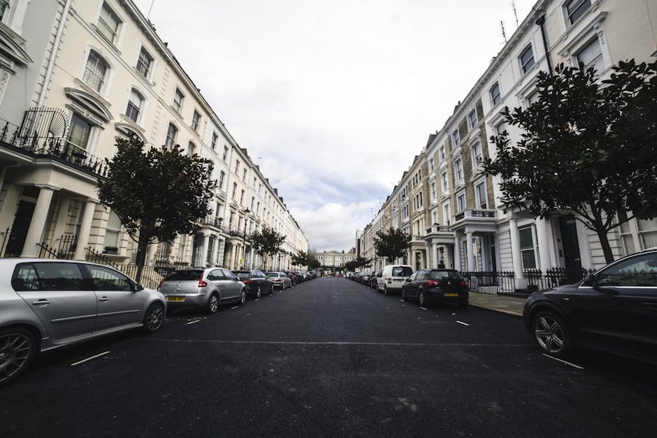 A wide residential street with white terraced houses featuring decorative balconies and bay windows on both sides. Several small trees line the pavement, and parked cars including hatchbacks, sedans,
