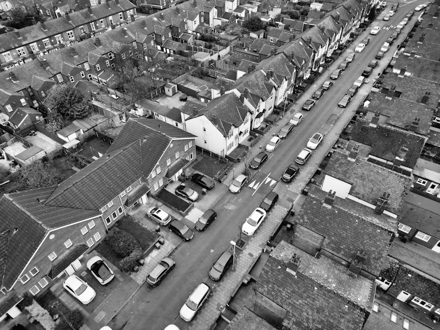 A wide residential street with white terraced houses featuring decorative balconies and bay windows on both sides. Several small trees line the pavement, and parked cars including hatchbacks, sedans,
